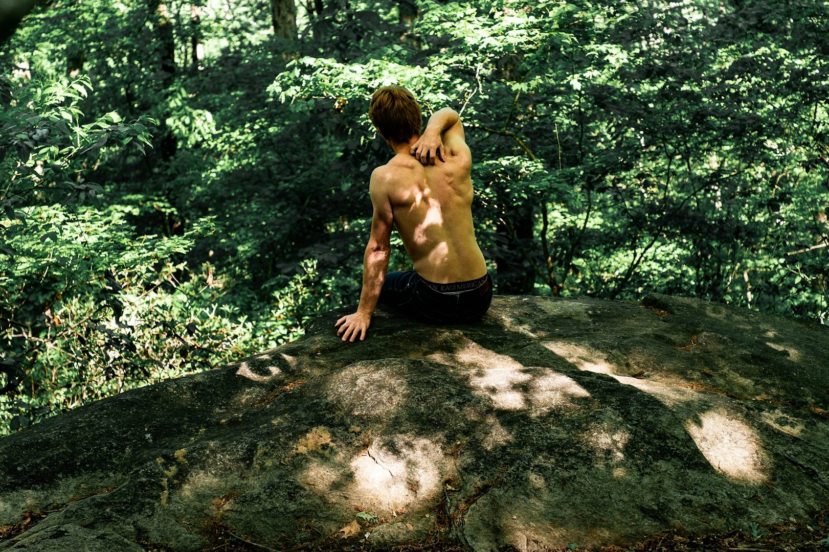 Stressmedizin Mann mit freiem Oberkörper sitzt auf einem Felsen im Wald, umgeben von Bäumen.